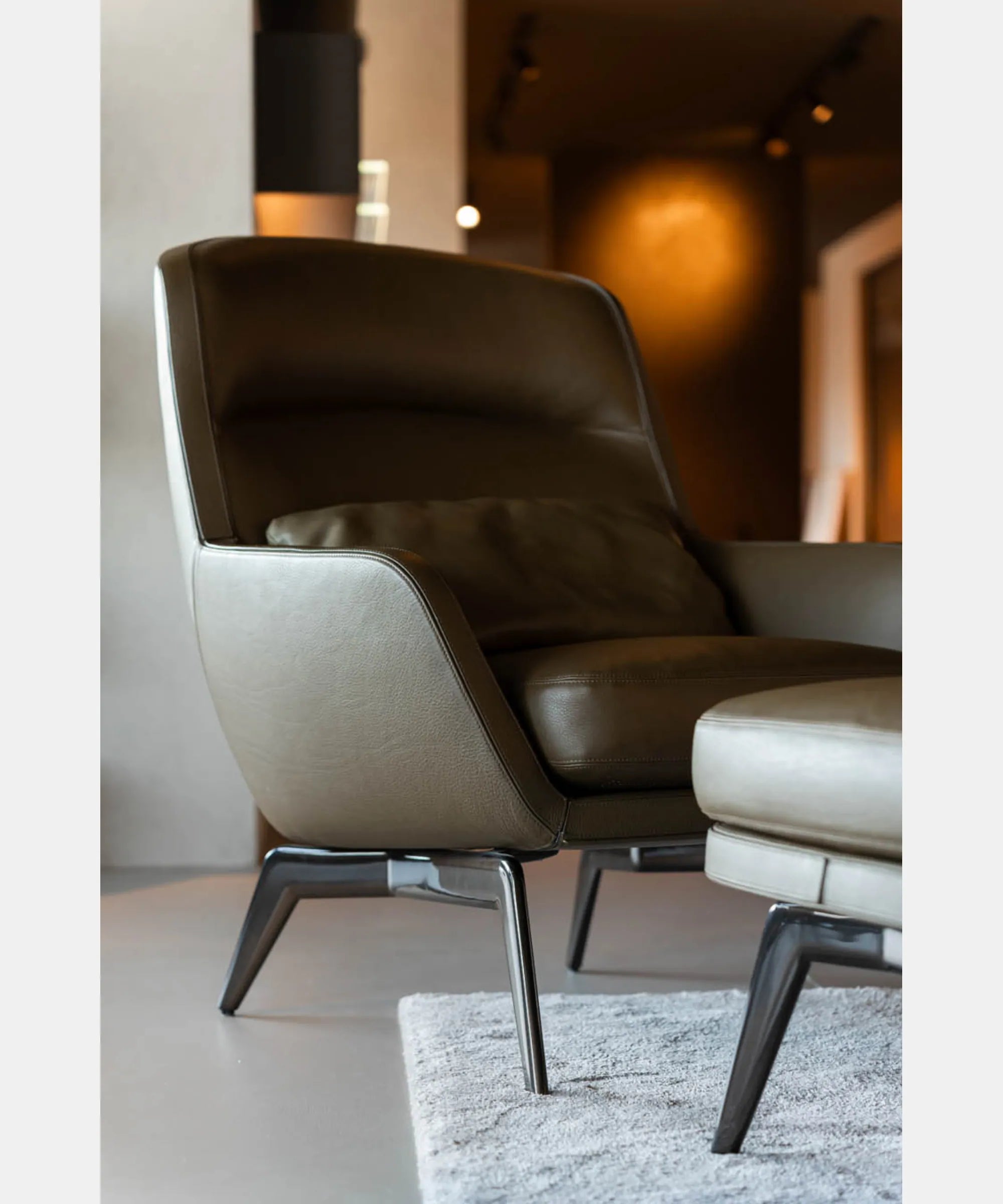 Poltrona Minotti Belt con poggiapiedi coordinato inserita in un ambiente contemporaneo, caratterizzata da rivestimento in pelle color, cuscino decorativo e gambe in metallo scuro.